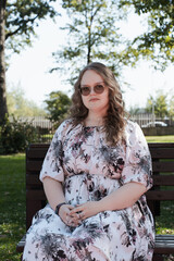 Young woman in a floral dress and sunglasses sitting on a park bench on a sunny day, looking at the...