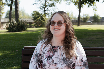 Young woman in a floral dress and sunglasses sitting on a park bench on a sunny day, looking at the...
