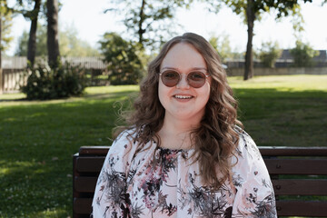 Young woman in a floral dress and sunglasses sitting on a park bench on a sunny day, looking at the...