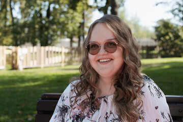 Young woman in a floral dress and sunglasses sitting on a park bench on a sunny day, looking at the...