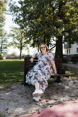Young woman in a floral dress and sunglasses sitting on a park bench on a sunny day, looking at the...