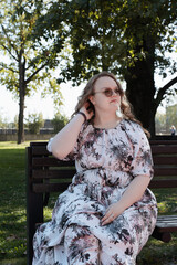 Young woman in a floral dress and sunglasses sitting on a park bench on a sunny day.