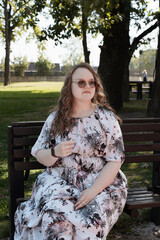 Young woman in a floral dress and sunglasses sitting on a park bench on a sunny day.