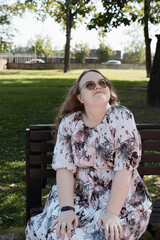 Young woman in a floral dress and sunglasses sitting on a park bench on a sunny day.
