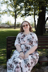 Young woman in a floral dress and sunglasses sitting on a park bench on a sunny day, looking at the...