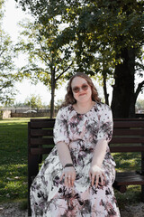 Young woman in a floral dress and sunglasses sitting on a park bench on a sunny day, looking at the...