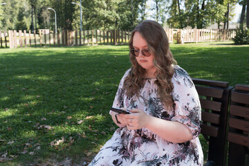Young woman with long curly hair using a smartphone outdoors in a park on a sunny day, smiling and...