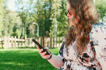 Young woman with long curly hair using a smartphone outdoors in a park on a sunny day, smiling and...