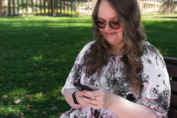 Young woman with long curly hair using a smartphone outdoors in a park on a sunny day, smiling and...