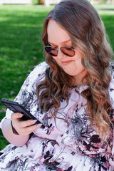 Young woman with long curly hair using a smartphone outdoors in a park on a sunny day, smiling and...