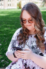 Young woman with long curly hair using a smartphone outdoors in a park on a sunny day, smiling and relaxed.