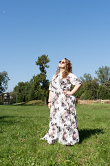 A young woman in a floral dress and sunglasses is posing joyfully on a grassy field in the park on...