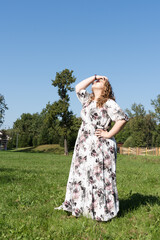 A young woman in a floral dress and sunglasses is posing joyfully on a grassy field in the park on...
