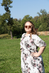 A young woman in a floral dress and sunglasses is posing joyfully on a grassy field in the park on a sunny day