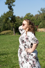 A young woman in a floral dress and sunglasses is posing joyfully on a grassy field in the park on a sunny day