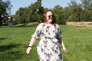 A young woman in a floral dress and sunglasses is posing joyfully on a grassy field in the park on a sunny day