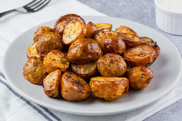 Golden brown roasted baby potatoes arranged neatly on a plate beside a small bowl of creamy dipping sauce, perfect for a savory snack or side dish. Rustic homemade food on a baking tray.