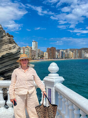 Woman in light-colored linen outfit stands by the ocean, holding a stylish bag, with a scenic...