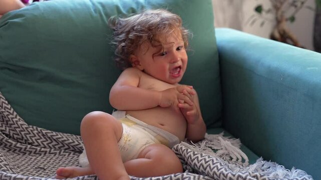 Smiling toddler lounges on teal couch wrapped in patterned blanket, looking off to side with playful expression in cozy indoor setting