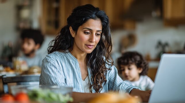 Young indian woman wife and mother remote working online from home office while family spending time at home. Busy parent mom using laptop computer sitting at table in homeoffice., no logos, no brand