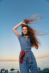 modern boho-western denim street style woman smiling with long hair outdoors by the sea at sunset,...