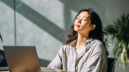 Young asian woman relaxes at office desk with laptop. Enjoying break moment, resting after work during working hours. Concept of workplace relaxation, mental health, stress relief, comfort lifestyle.