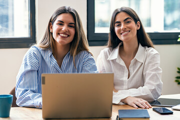 Two beautiful designer women working together with laptop while looking at camera in the office