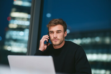 Focused young businessman working late at night in office with laptop and mobile phone