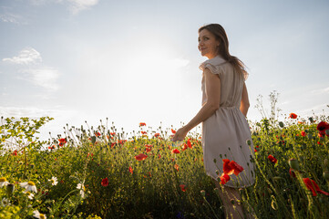 Young woman in a light summer dress walking through a poppy field at sunset. Peaceful countryside...
