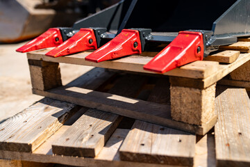 Excavator bucket teeth with bright red tips resting on wooden pallets at construction site with...