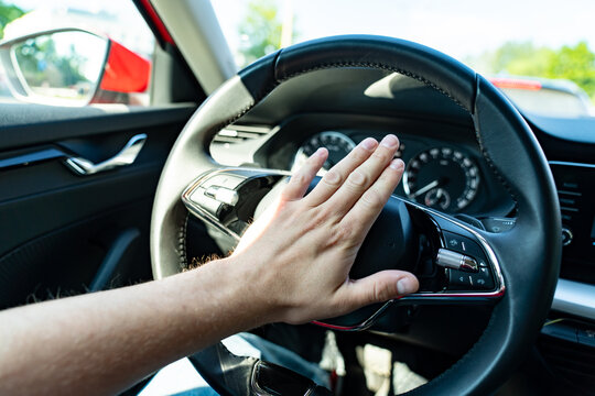 Driver hand pressing car horn button on steering wheel inside modern vehicle showing dashboard, speedometer and car interior details during daytime traffic situation