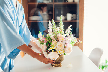 Woman puts brass vase with bouquet full of roses flowers on table. Interior and home decor. Summer...