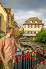 A man stands on a bridge adorned with love locks in La Petite Venise, Colmar, overlooking the Lauch river and picturesque Alsatian houses—ideal for travel, romance and leisure concepts