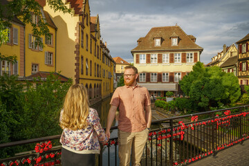 A loving couple stands on a bridge adorned with love locks in La Petite Venise, Colmar, overlooking the Lauch river and charming Alsatian houses—ideal for romance, travel, and leisure concepts.