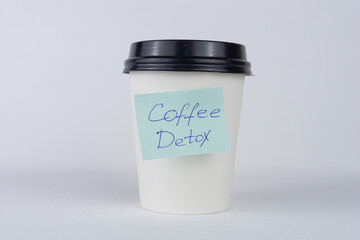 A white disposable coffee cup with a black lid and a light blue sticky note on it, handwritten with...