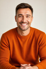 Obraz premium A smiling man with short brown hair and a light beard gazes directly at the camera, wearing an orange sweater against a soft gray background.