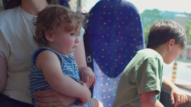 Baby sitting upright in mother&rsquo;s arms looking out on bus while older child leans against window, both watching passing city scenery