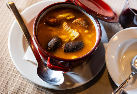 In deep baking dish there is portion of Asturian fabada - thick soup of beans, smoked bacon and spices. Ladle and small amount of alcoholic aperitif are served with dish