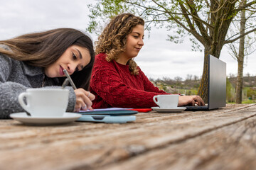Two young female students studying outdoors at a wooden picnic table with coffee and laptop