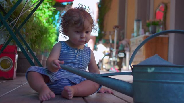 Toddler plays with watering can while sitting on wooden balcony floor, exploring quietly in striped romper as soft light filters through plants around him