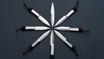 Top-down view of a set of steel throwing knives with cord-wrapped handles arranged in a circular star pattern on a dark slate background.
