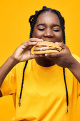 Happy young woman enjoying a delicious burger against a bright yellow background, showcasing her...