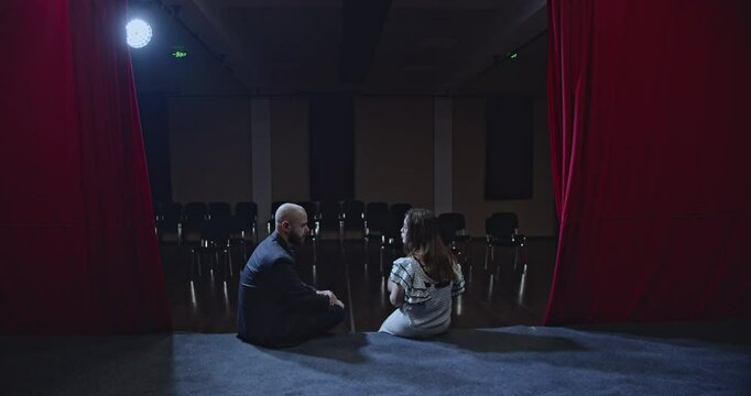 Two young actors sit on the edge of a stage in an empty auditorium, discussing their roles and performance.