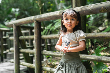 Curious Latina toddler is standing by a wooden zoo railing gazing into the camera amid lush greenery