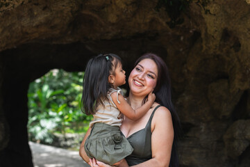 Smiling mother holds her young Latina daughter kissing inside a shady zoo cave surrounded by foliage