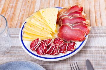 On plainly set table - plate with hearty meat snacks and yellow hard pressed cheese. Circles of...