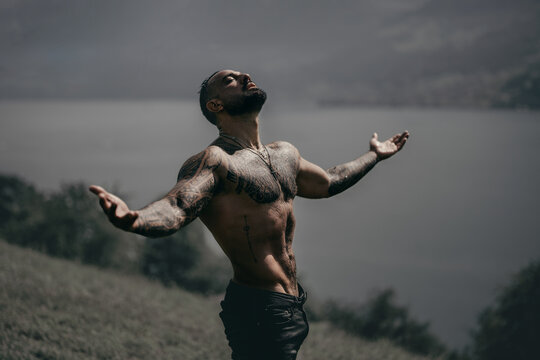 Carefree man in nature. Freedom man in alpine landscape. Muscular model breathing air near waterfall. Handsome model with raised hands outdoors. Fit lifestyle. Relaxed man. Sexy man. Healthy torso.