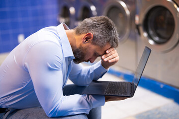 Man handling housework and workload. Man doing laundry and working on laptop. Man multitasking...
