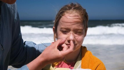 A mother applies sunscreen to her child face at the beach on a sunny day. The close-up captures a caring moment of sun protection near the ocean waves and beach. - Powered by Adobe
