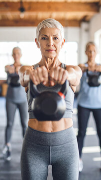 Personas mayores levantando pesas tipo kettlebell en una clase grupal de ejercicio con fondo claro y ambiente de fuerza y vitalidad